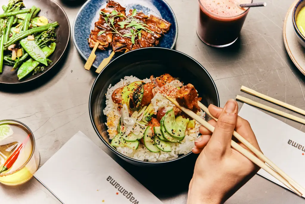 hands holding chopsticks with wagamama dishes and drinks in front of them